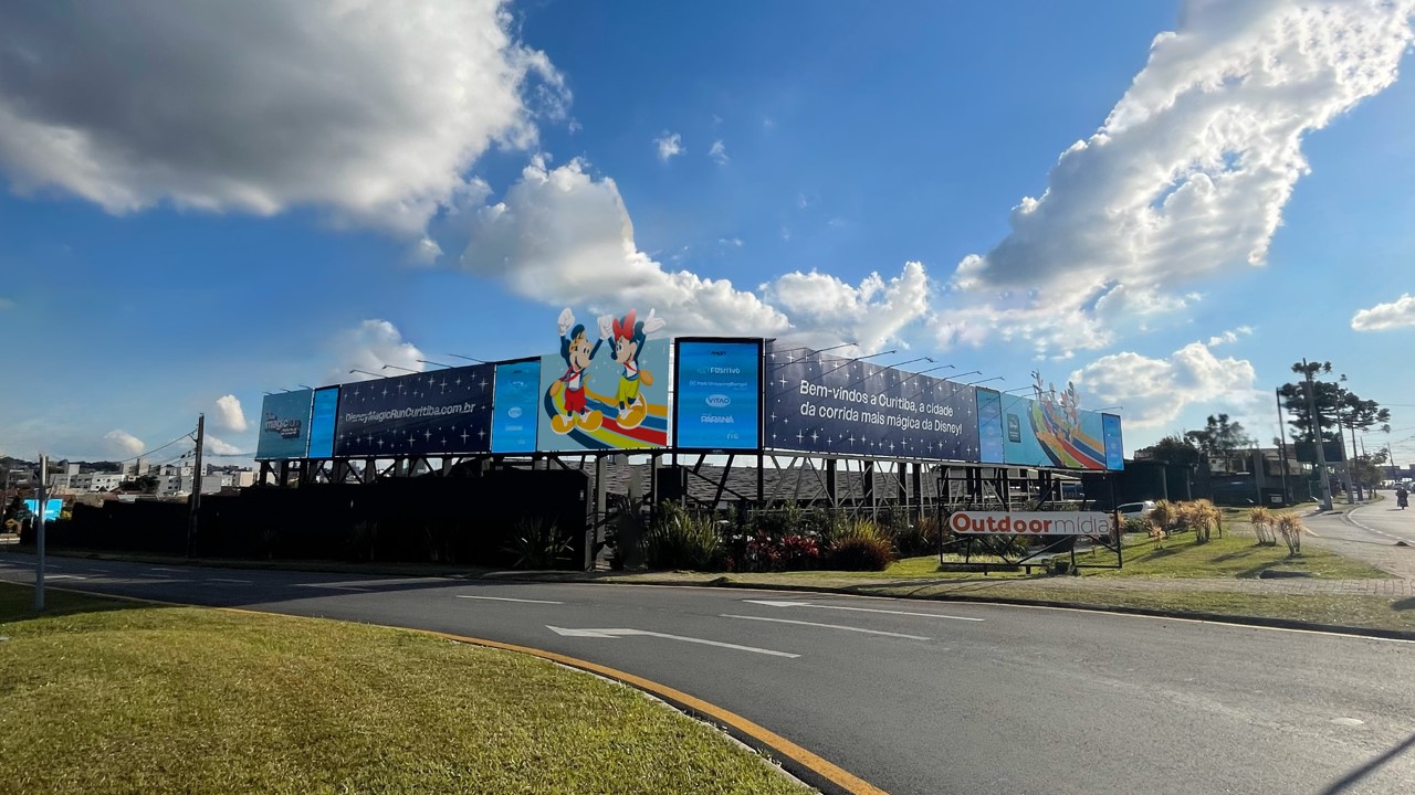Sequência de painéis exibindo uma campanha automotiva na via de saída do aeroporto, sob céu azul
