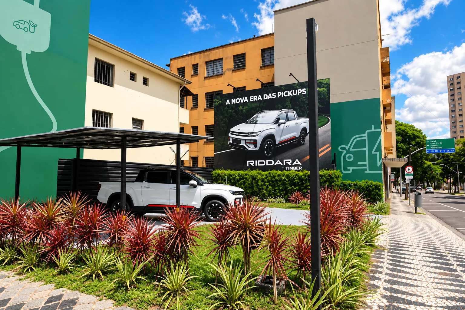 Painel com campanha da Riddara ao lado da cobertura solar dos carregadores, com uma picape branca estacionada e canteiro de plantas na esquina