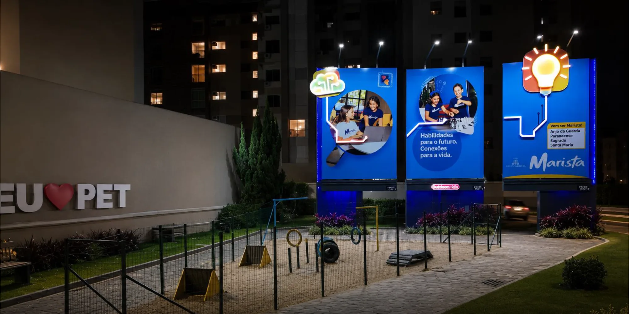 Praça pet iluminada à noite, com letreiro, brinquedos e painéis do patrocinador