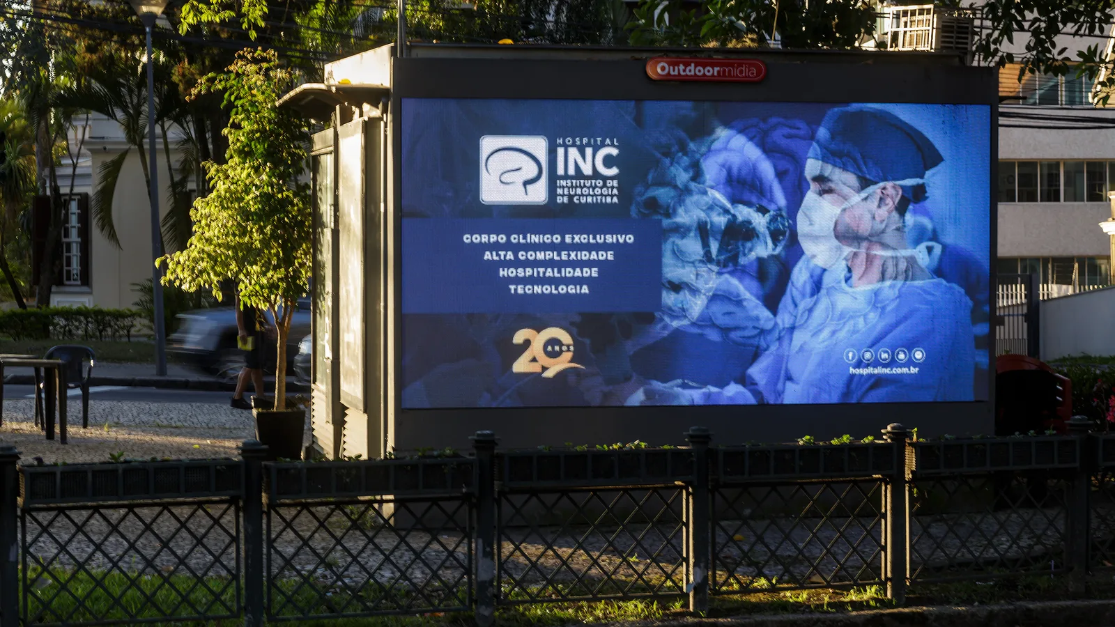 Banca digital da Outdoormídia em calçada arborizada de Curitiba, tela de LED com anúncio do Hospital INC.