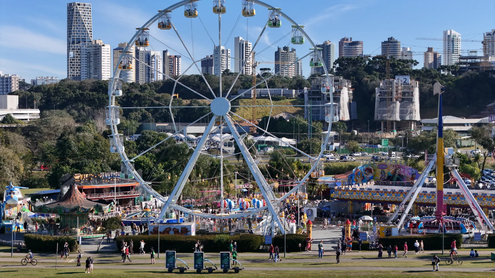 Trio de Bike Mídia da Outdoormídia circulando no gramado de um parque de Curitiba, com a roda-gigante ao fundo