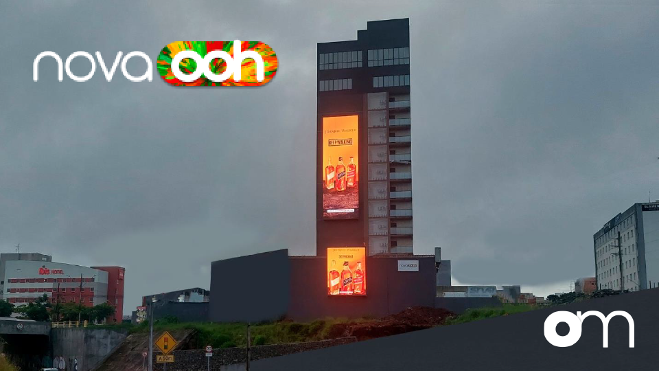 Empena digital da Outdoormídia na lateral de um edifício ao entardecer, com dois painéis de LED acesos exibindo a mesma campanha