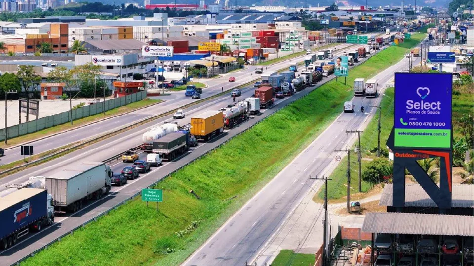 Painel Urbanity com campanha da Select Planos de Saúde às margens da rodovia, com a placa para Itajaí e Balneário Camboriú e a fila de caminhões na pista